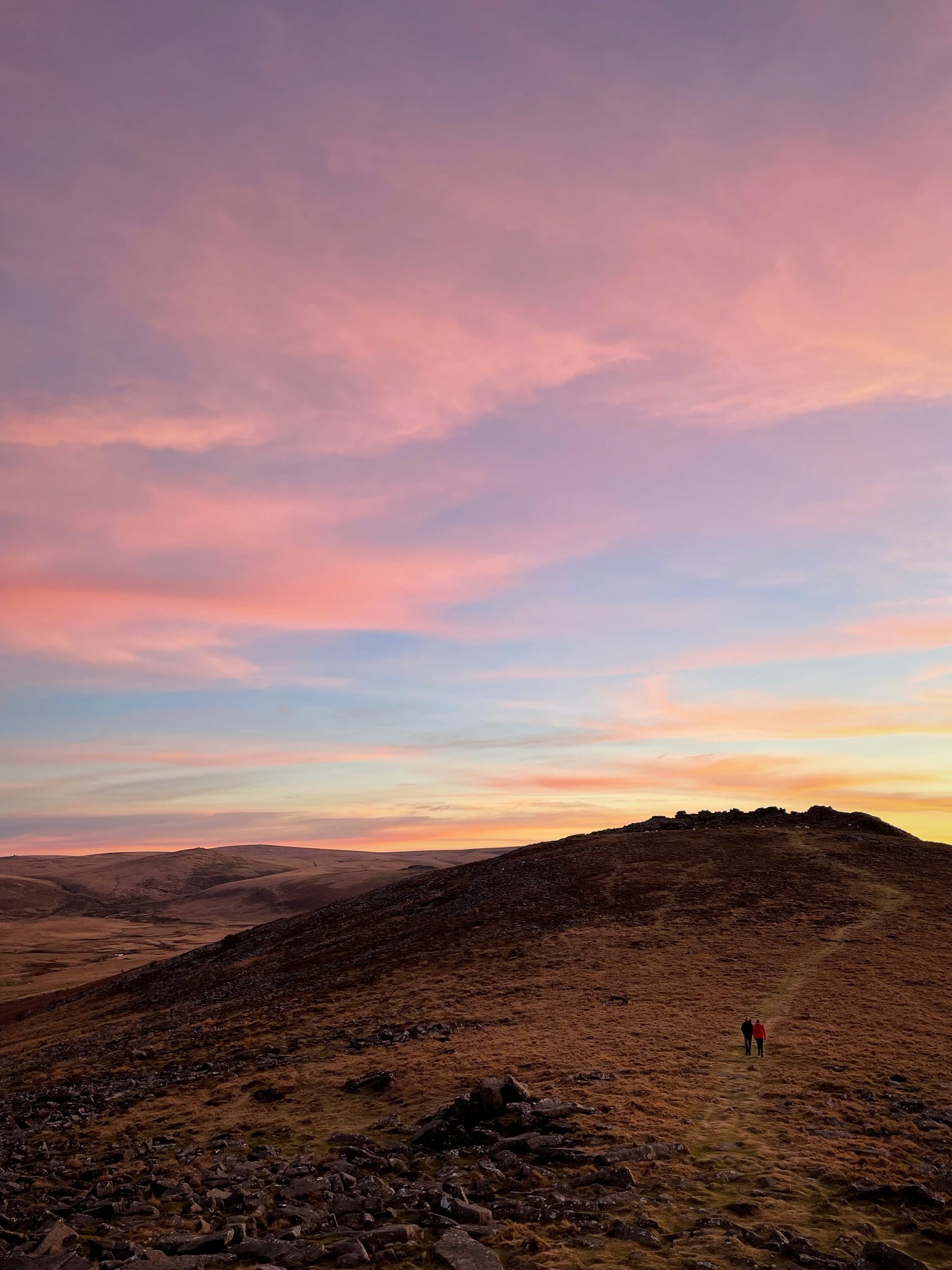 two people walking below a Pink and orange sunset sky over Higher Tor, Belstone, Dartmoor, Devon