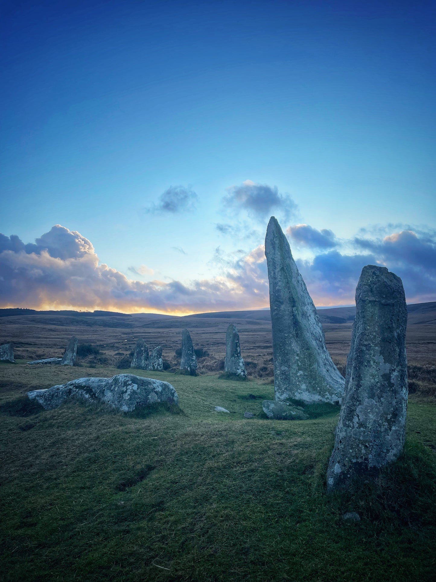 Scorhill Stone Circle at blue hour, Dartmoor -- ancient granite standing stones silhouetted against a glowing twilight sky of deep blue and warm amber, with open moorland stretching to the horizon.